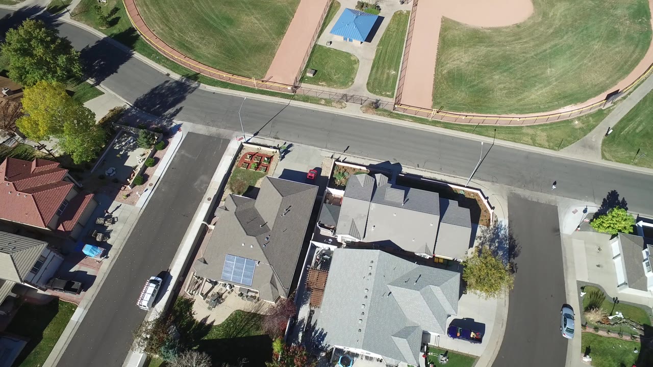 Overhead frame in Hidden Lake, Colorado showing rooftops, a curved residential street, a cross street, trimmed lawns, and the green outfield across the road.
