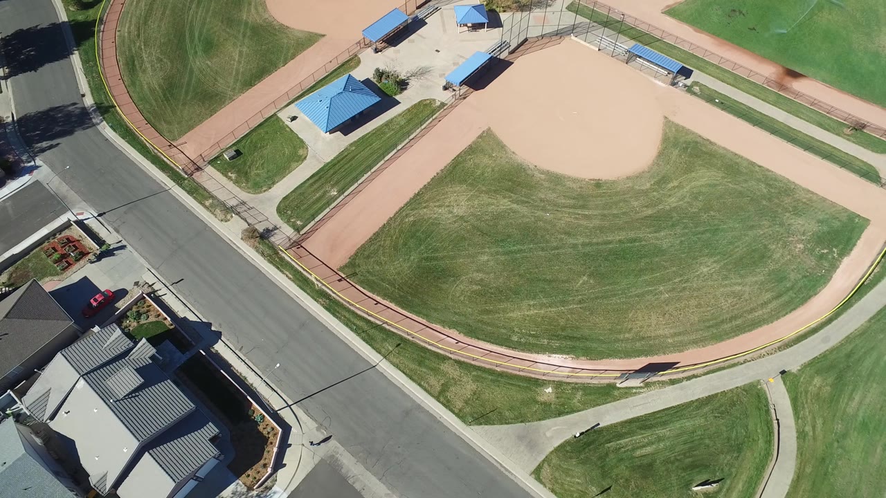 Aerial view over Hidden Lake, Colorado showing the baseball diamond shifted slightly across frame, with the road at left, homes below, blue-roofed park shelters, and no visible people.