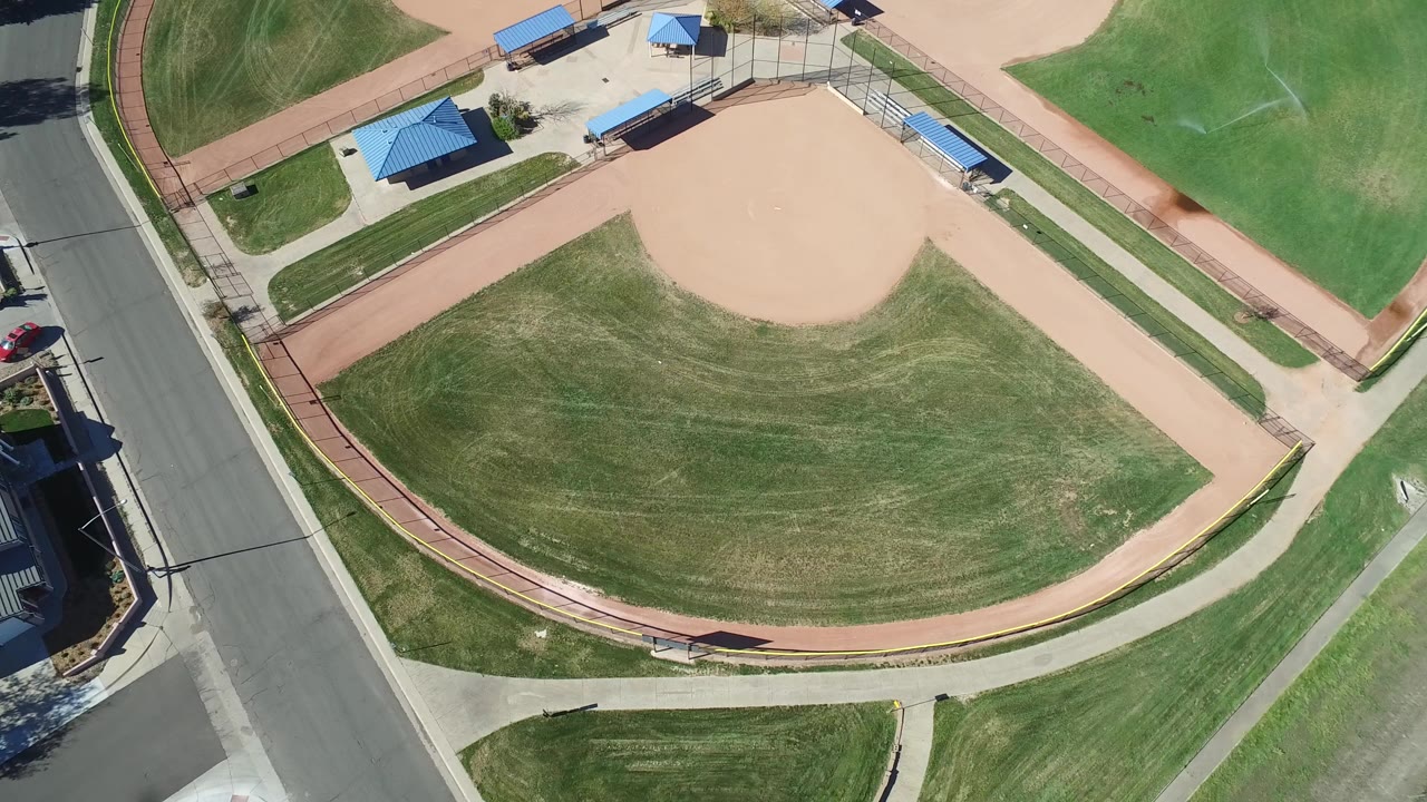 Overhead view of a baseball field beside a road and blue-roof shelters.