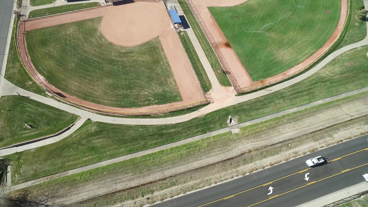 Aerial view in Hidden Lake, Colorado showing a road at the bottom edge, sidewalks, mowed grass, baseball infields, and no visible people.