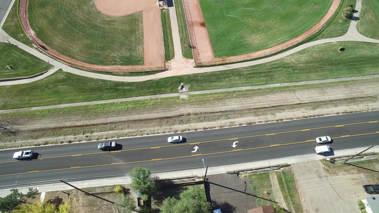 Overhead road beside green baseball fields with several cars.