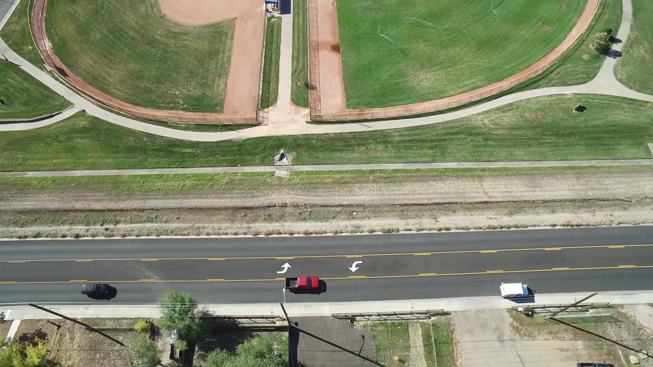 Overhead aerial frame in Hidden Lake, Colorado showing green sports fields, tan dirt infields, a concrete path, a road with several cars, and no visible people.