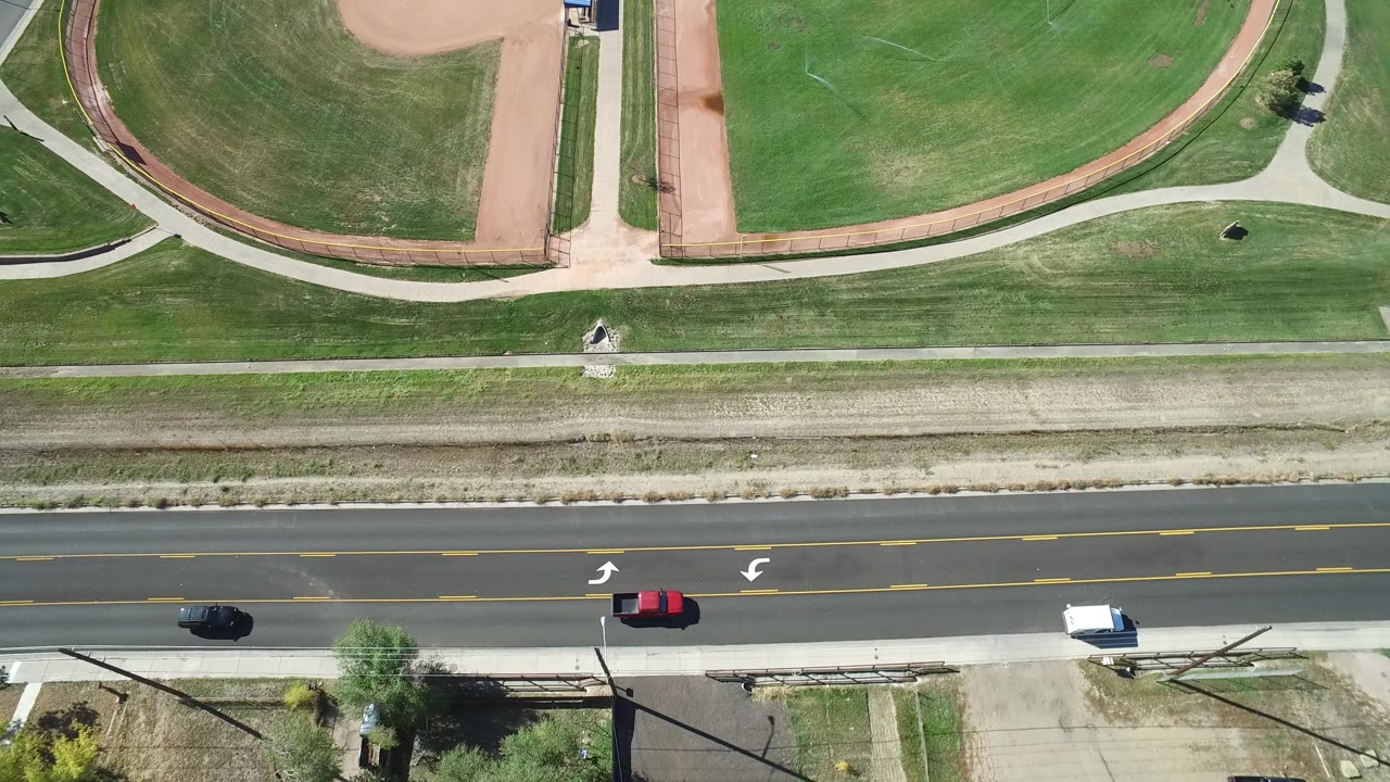 Aerial frame in Hidden Lake, Colorado with curved ball-field dirt, bright green grass, a middle walking path, a paved road, moving vehicles, and no visible people.