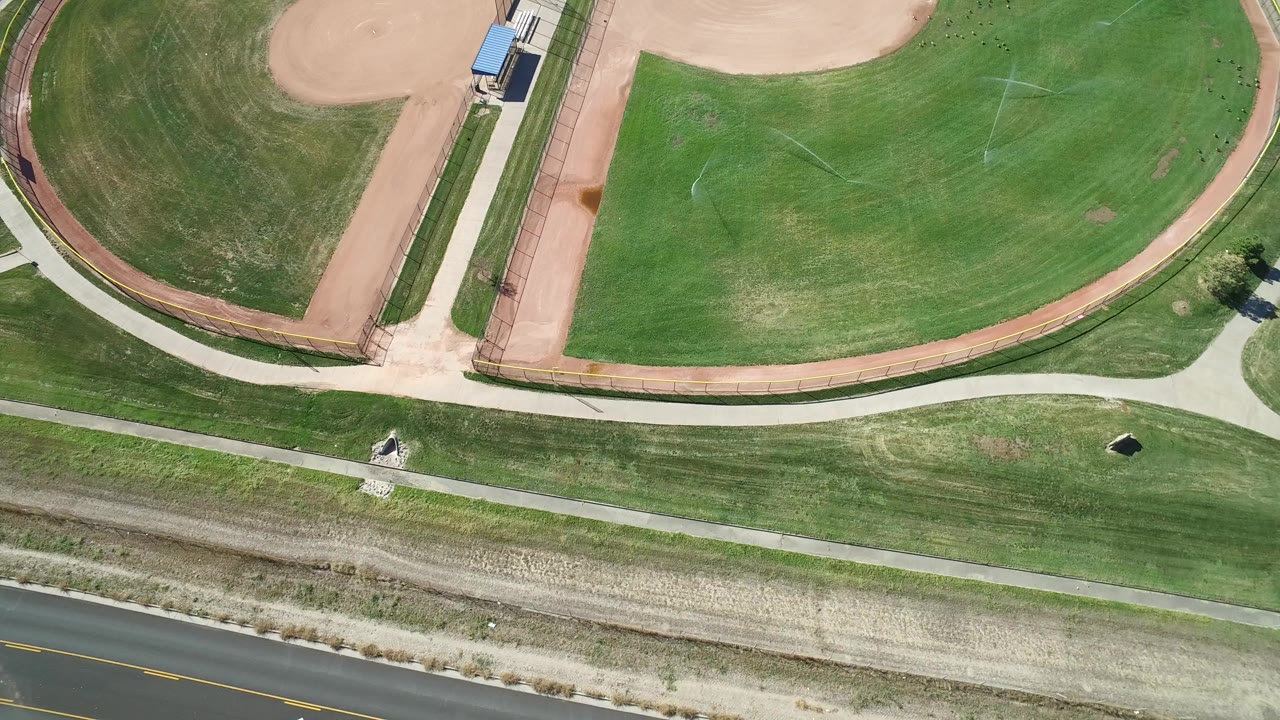 Overhead frame from Hidden Lake, Colorado showing green ballfields, tan infield dirt, paved paths, a road along the bottom edge, and no visible people.