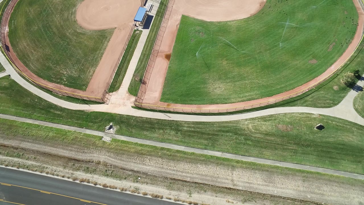 Aerial frame from Hidden Lake, Colorado with two tan baseball infield areas, a broad green outfield, curving pedestrian paths, roadside pavement, and no visible people.