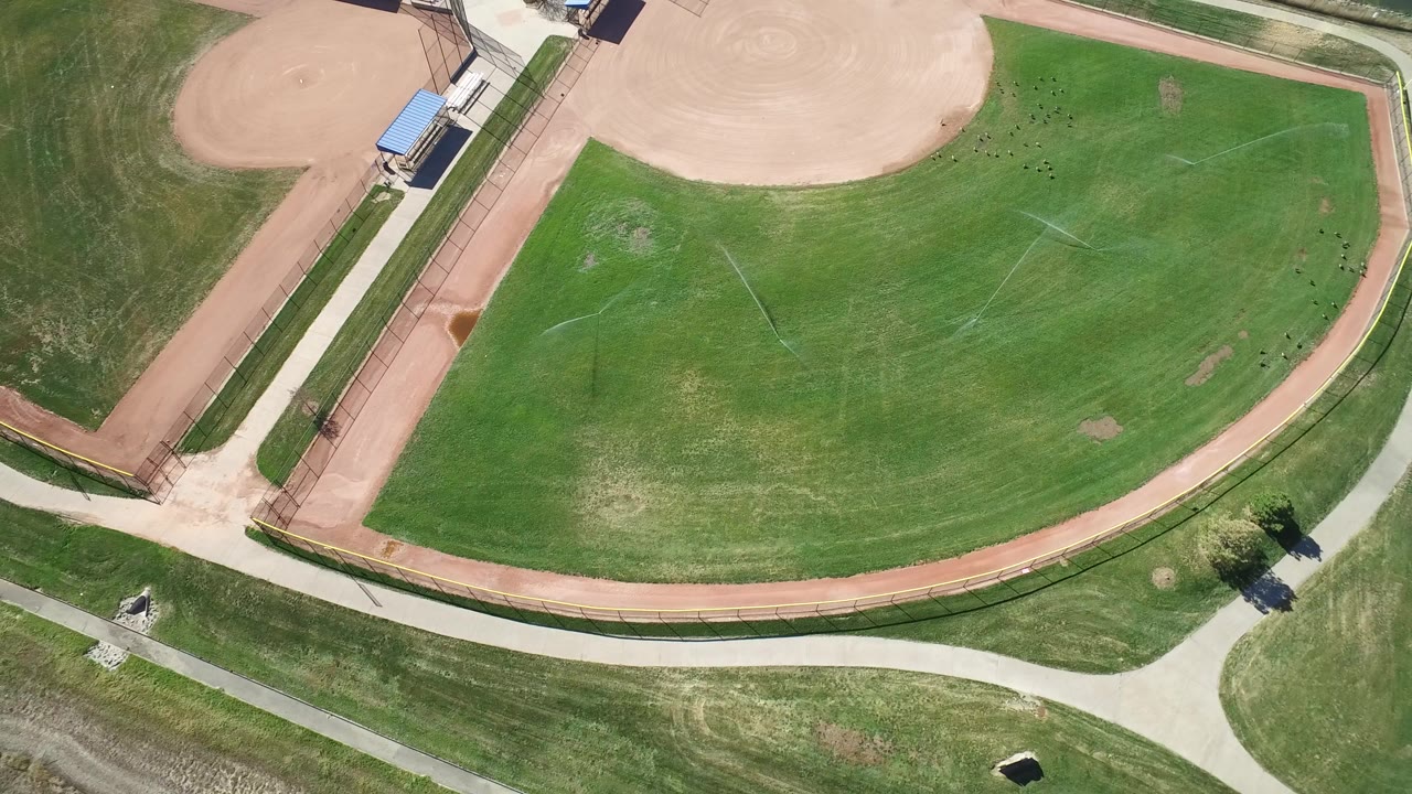 Overhead view in Hidden Lake, Colorado showing a green baseball outfield, tan infield edge, paved paths, a bordering waterway, and no visible people.