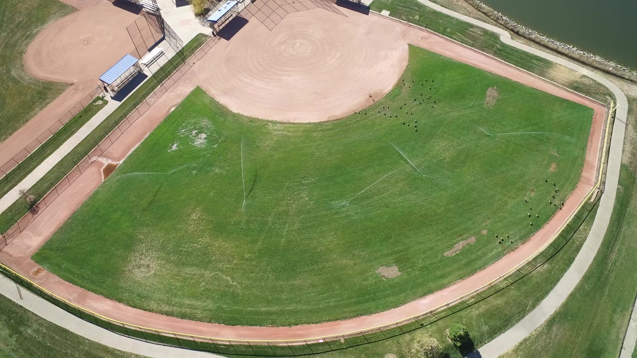 High overhead frame in Hidden Lake, Colorado showing a green recreation field, red perimeter track or path, tan baseball infield, small structures, lake water at the upper right, and no visible people.