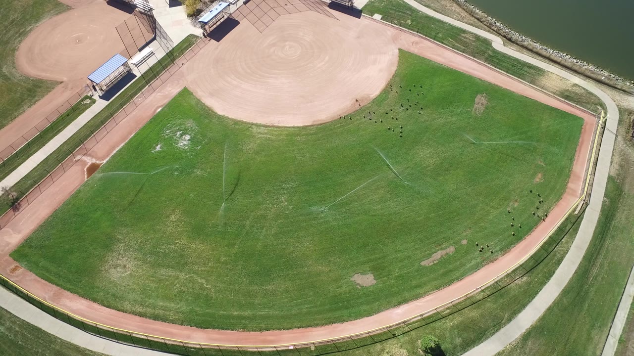 Top-down view of an empty field with baseball dirt, walkways, and lake water nearby.