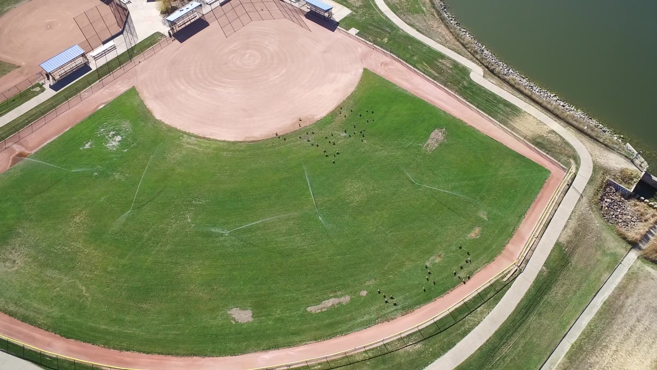 Overhead drone-style view in Hidden Lake, Colorado, showing a tan baseball infield, green outfield, lakeside path, rock shoreline, and no visible people.