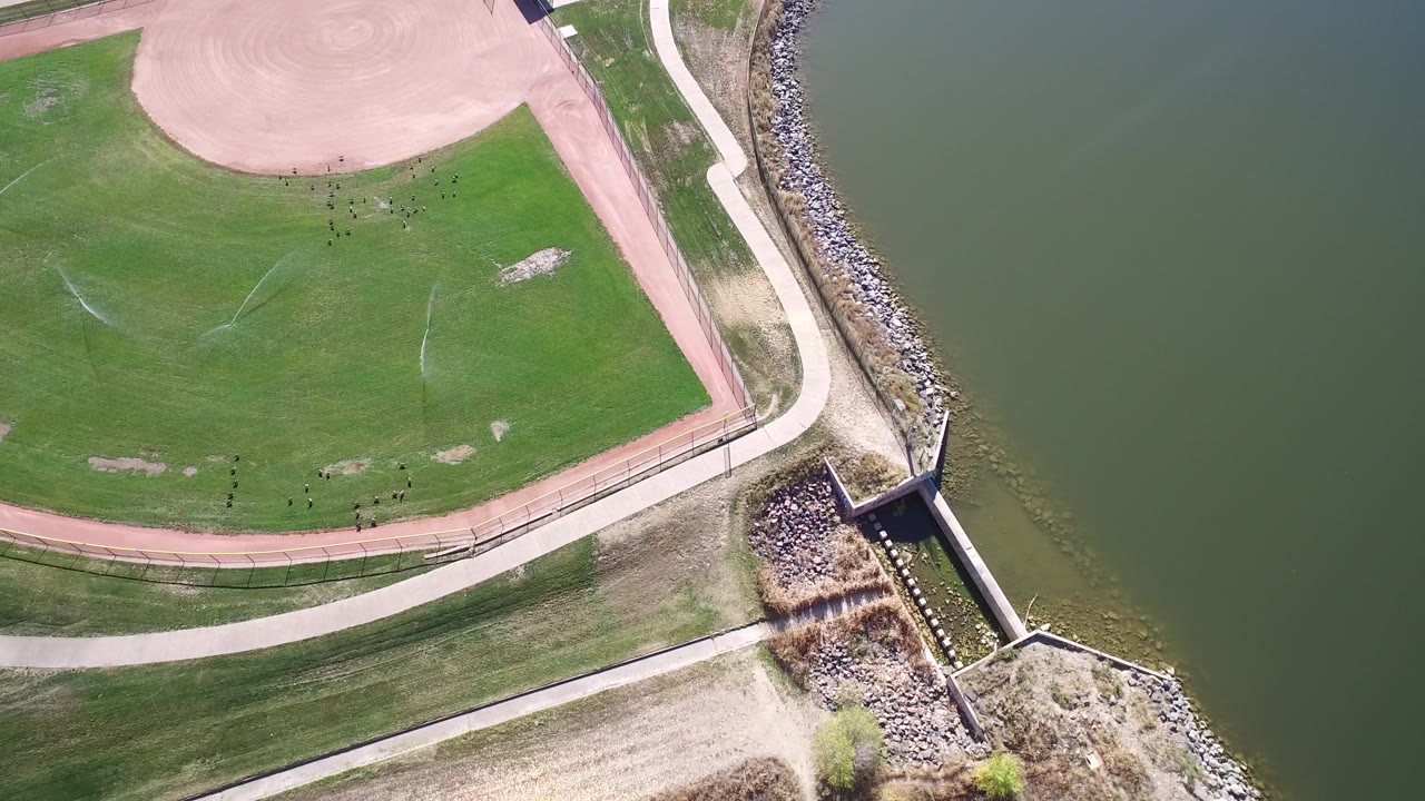 Overhead drone frame in Hidden Lake, Colorado showing a green sports field, reddish track edge, pale paths, rock shoreline, and lake water with no visible people.