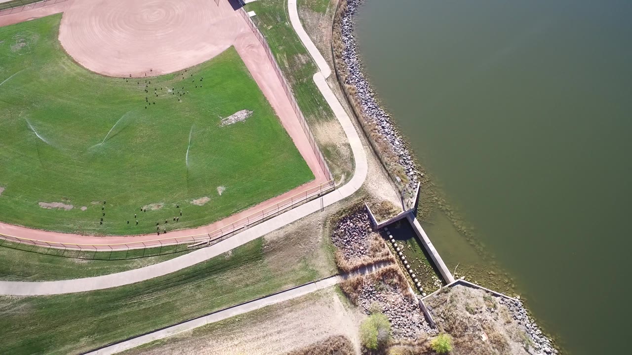 Hidden Lake, Colorado aerial frame with the sports field upper left, curved paths through grass, a rock-lined bank, a small shoreline structure, and open water at right with no visible people.