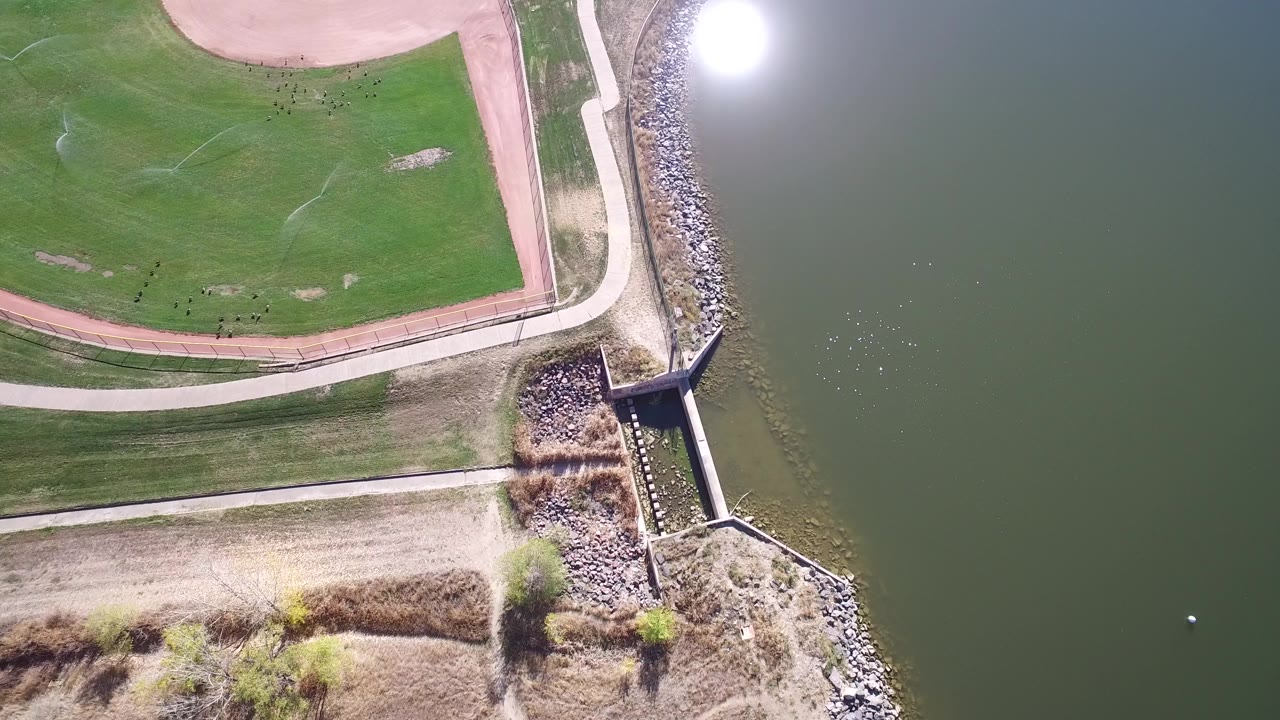 Overhead frame from Hidden Lake, Colorado showing green water, a rock-lined edge, paved paths, grass, bare ground, and no visible people.