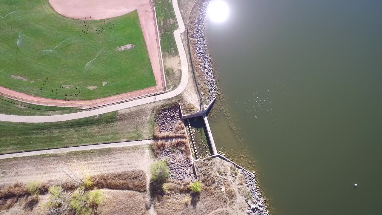 Aerial frame from Hidden Lake, Colorado with the water-control structure near the lower center, bright glare on the water, shoreline rocks, grass, and no visible people.