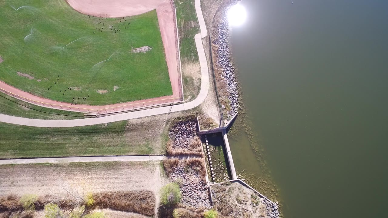 High overhead frame in Hidden Lake, Colorado with the green field and reddish infield edge at left, a pale path near the shore, riprap along the water, and no visible people.