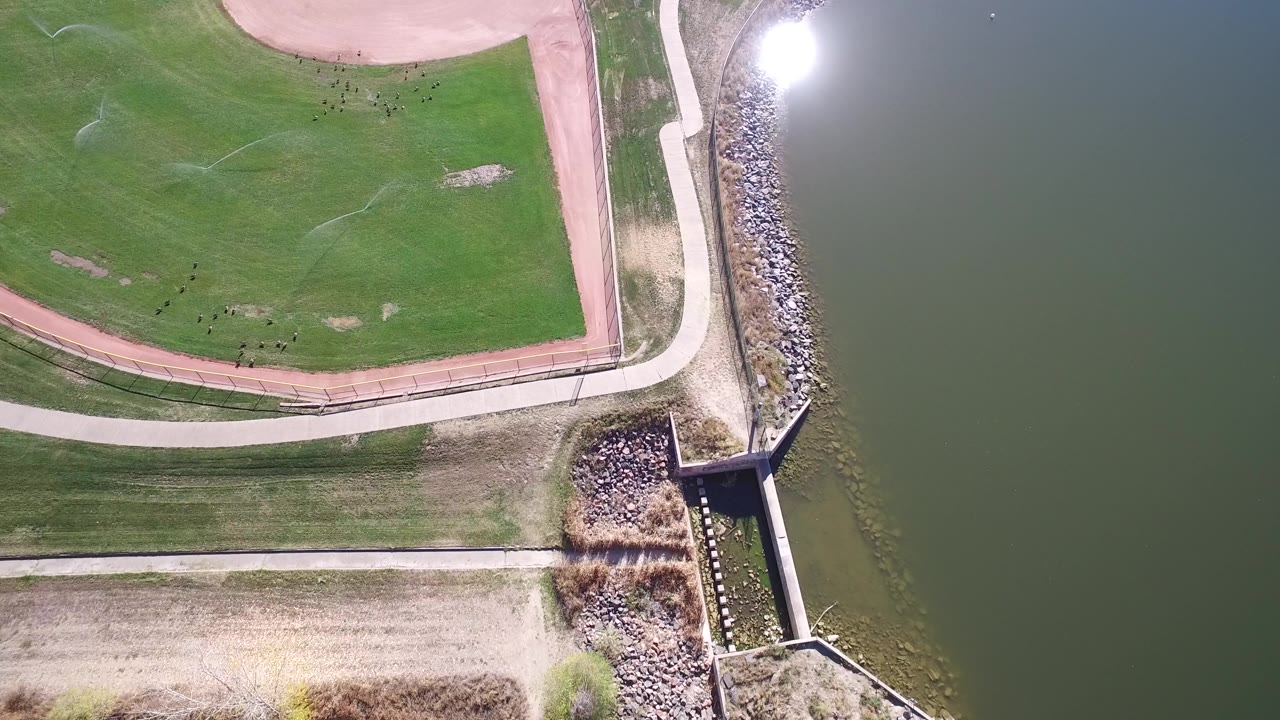 Top-down lakeside park with baseball field edge and bright water reflection.