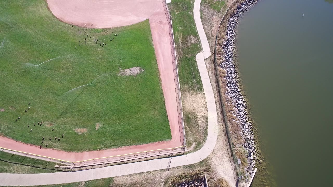 Opening overhead frame at Hidden Lake, Colorado, showing a green field bordered by a pink path, shoreline rocks, and dark water with no visible people.