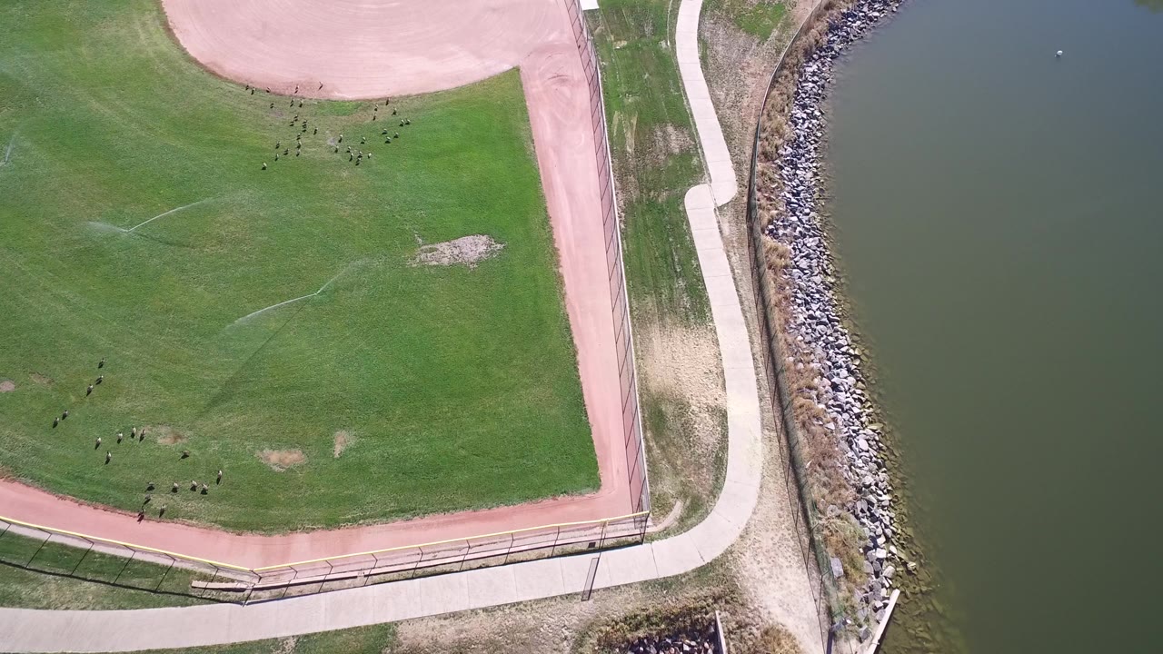 Second overhead frame at Hidden Lake, Colorado, with the grass field filling most of the view, the path curving around it, and the waterline tight along the right side with no visible people.