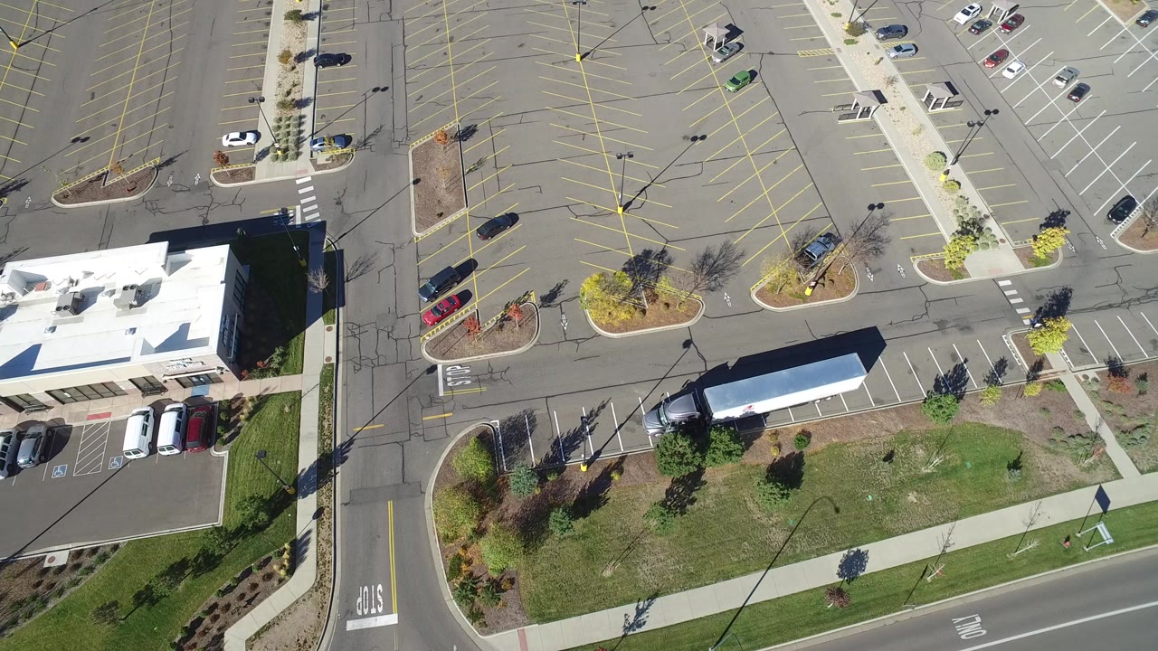 Opening overhead frame in Hidden Lake, Colorado showing a white-roofed low building, marked parking spaces, roads, sidewalks, grass, trees, parked cars, and no visible people.
