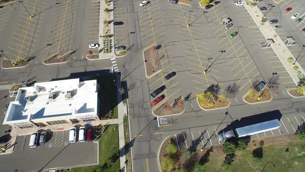 Drone-style overhead view in Hidden Lake, Colorado showing a white commercial building, a broad parking lot, small landscaped islands, parked vehicles, and no visible people.