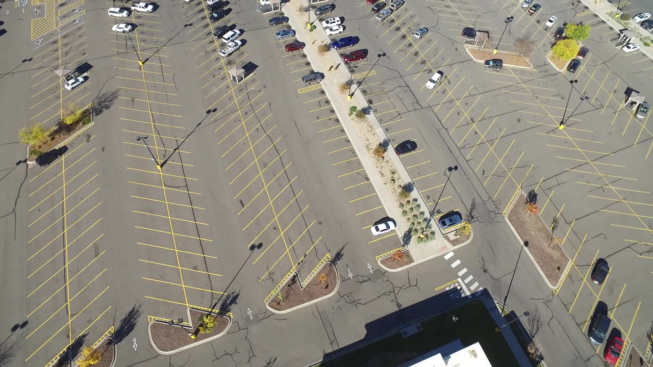 High aerial view over a Hidden Lake, Colorado parking lot with diagonal yellow stall lines, landscaped islands, light poles, a few cars, building edge shadows, and no visible people.