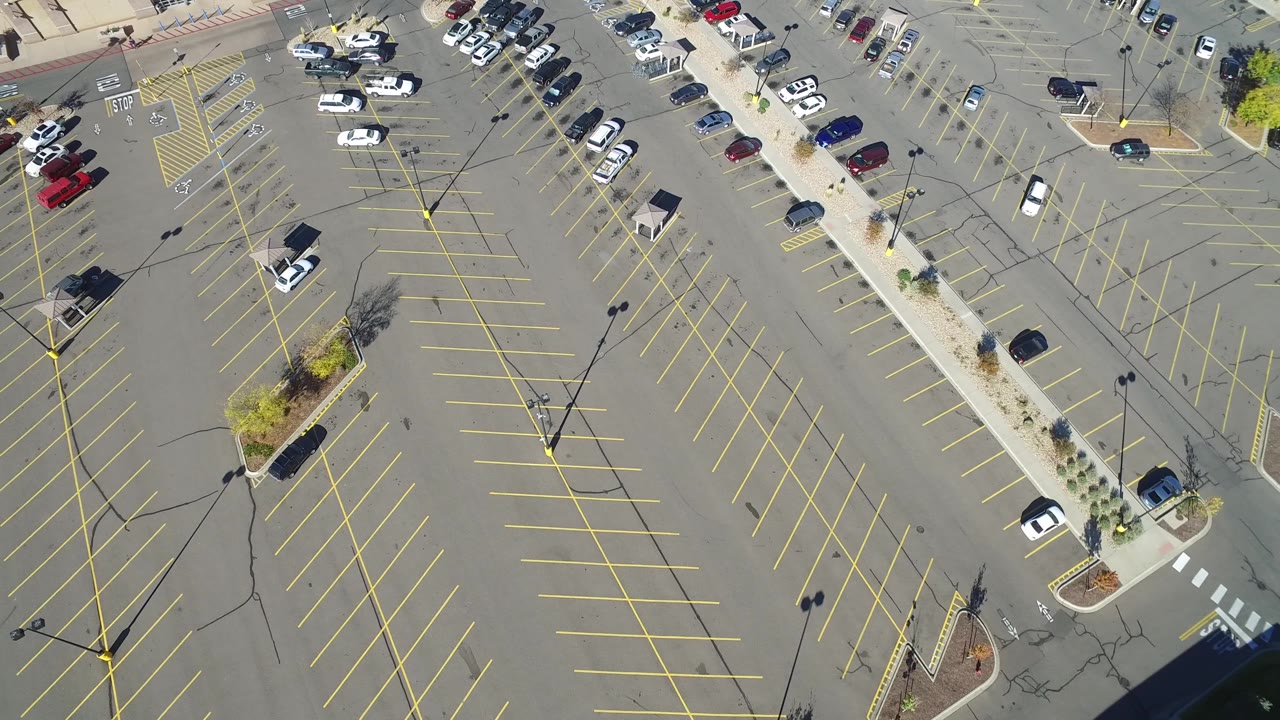 Aerial frame of the Hidden Lake parking area with empty yellow-striped rows across the center, cars gathered near the upper sidewalk, and a landscaped divider at left.
