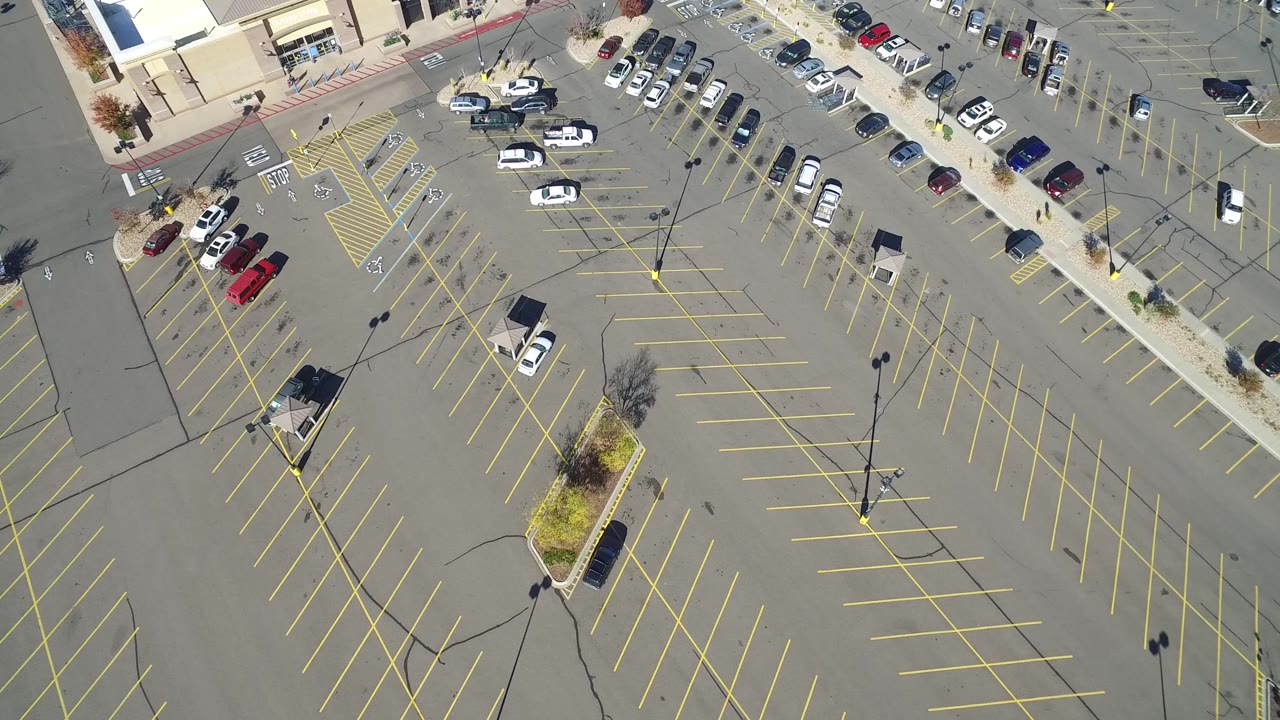 High overhead frame of the Hidden Lake, Colorado lot showing long rows of empty yellow-marked spaces, cars clustered by the sidewalk, planting islands, and no visible people.