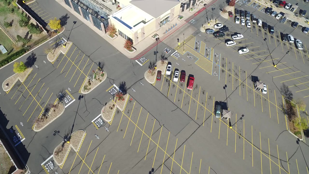 Overhead frame from Hidden Lake, Colorado showing a mostly empty parking lot, yellow stall lines, landscaped islands, parked cars near a building edge, and no visible people.
