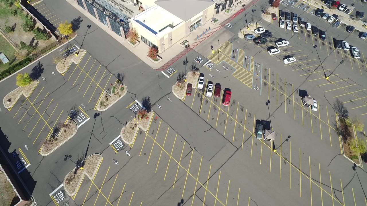 High aerial view in Hidden Lake, Colorado with empty yellow-lined parking rows, a cluster of parked cars near the building, landscaped medians, hard sunlight, and no visible people.
