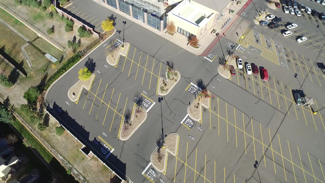 Overhead view of a commercial parking lot with yellow lines, landscaped islands, roads, cars, and a building edge.