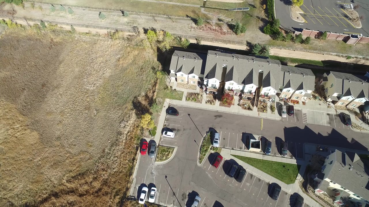 Aerial frame in Hidden Lake, Colorado with gray condo roofs along the right side, parked cars below, dry field on the left, roadway above, and no visible people.