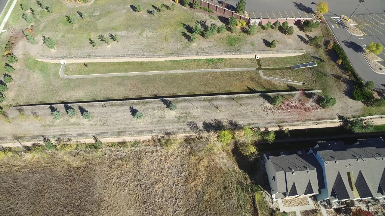 Overhead frame in Hidden Lake, Colorado showing pale open grassland with concrete trails, scattered trees, rooftops at the lower edge, a parking area at the top, and no visible people.