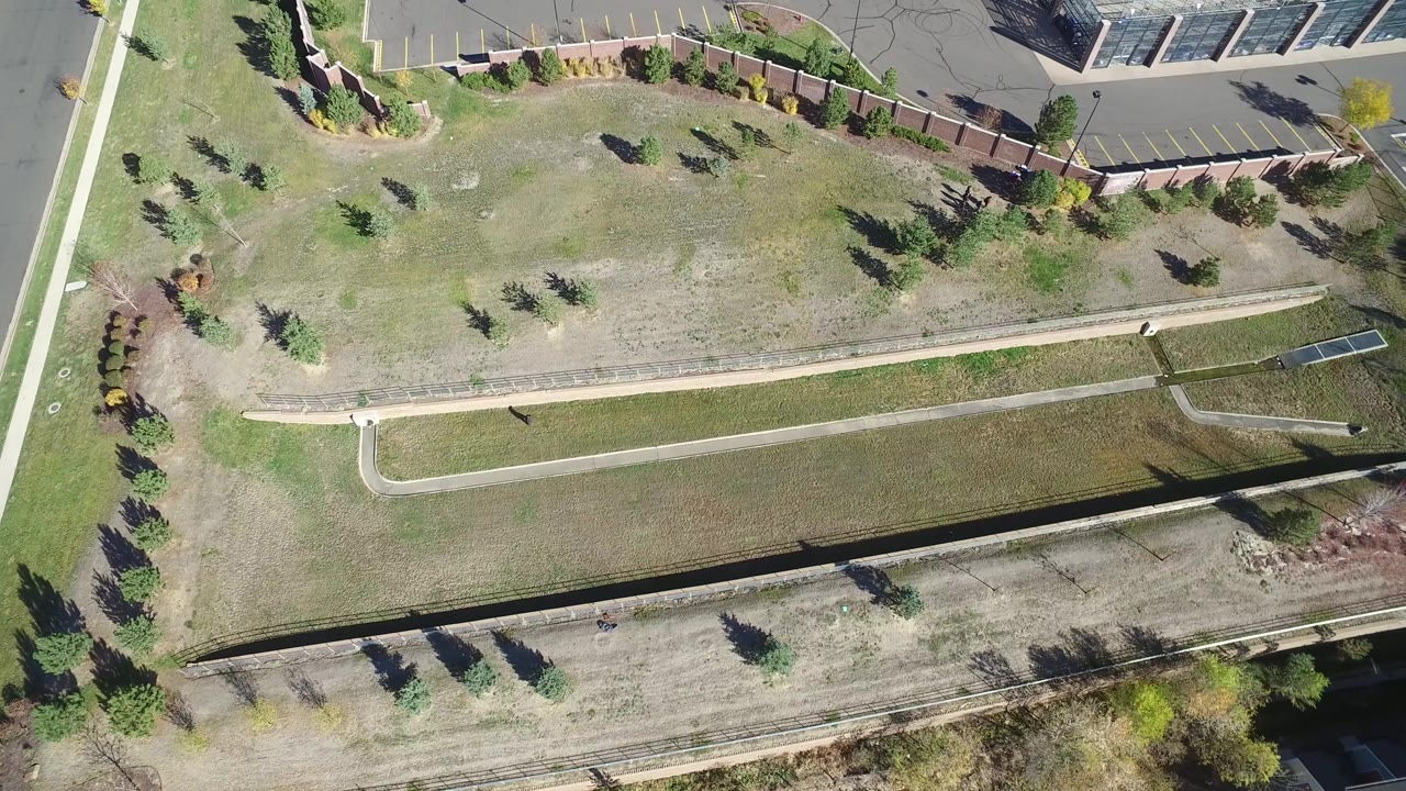 Top-down aerial frame in Hidden Lake, Colorado showing a dry grass field crossed by sidewalks, landscaped trees, a road edge, parking spaces, and building edges with no visible people.
