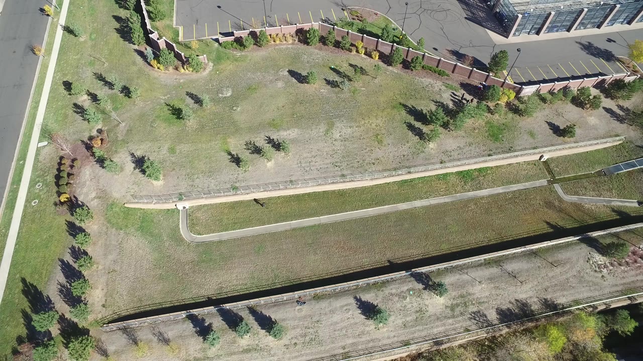 Drone view of open grass, curving paths, small trees, a road, and a parking lot corner.