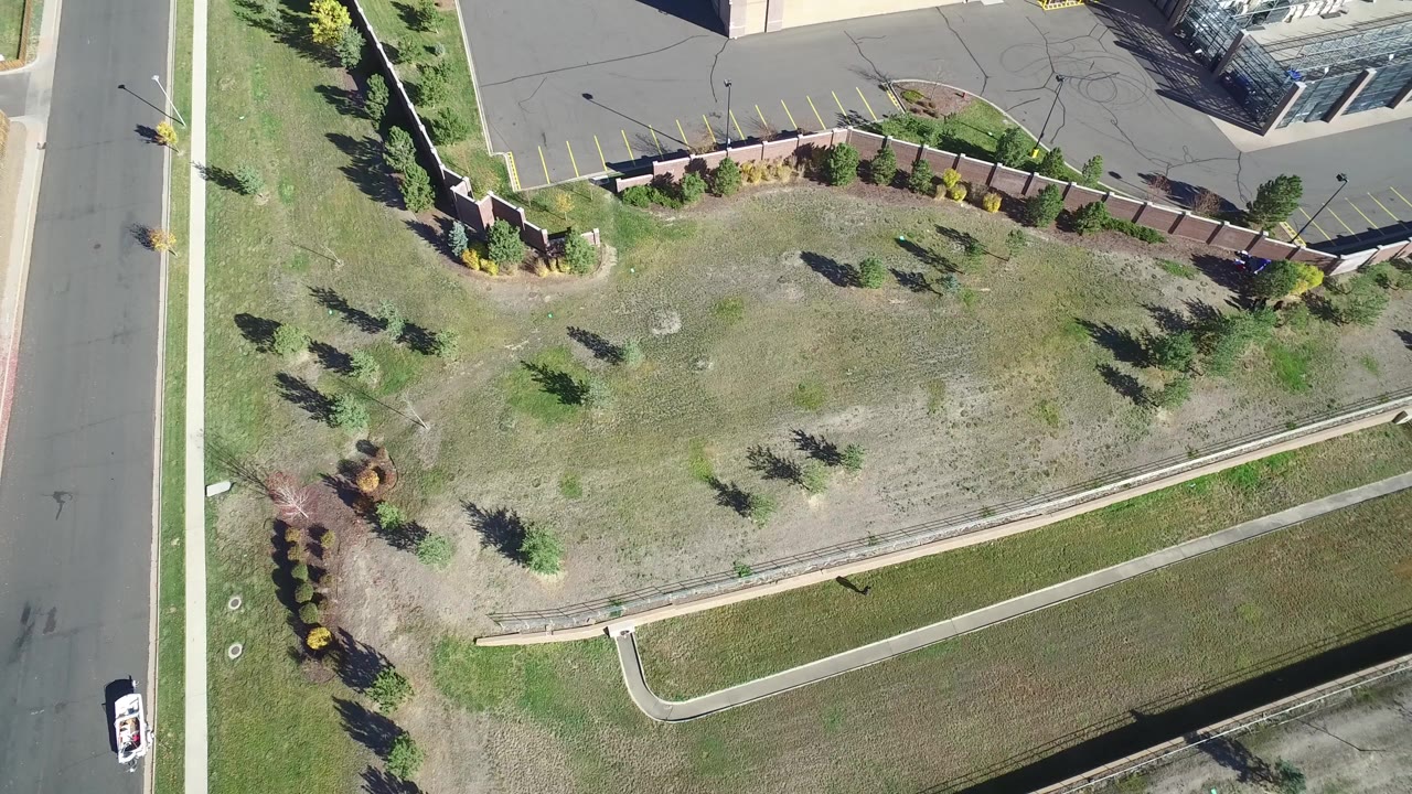 Overhead frame in Hidden Lake, Colorado showing a patchy green lawn beside a road, sidewalks, a curved wall, parking pavement, and building edges, with no visible people.