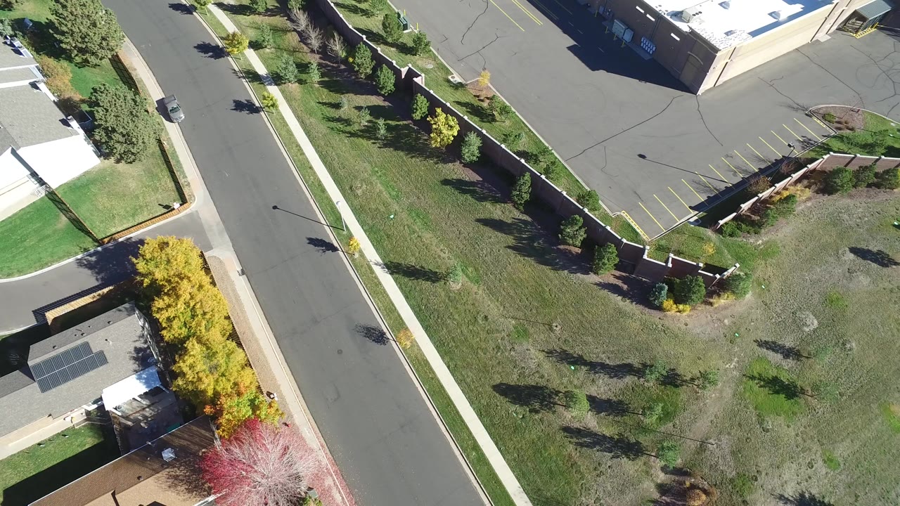 Opening overhead frame in Hidden Lake, Colorado, showing a diagonal paved road, sidewalks, green lawns, houses at the edges, trees, and a gray parking lot area.