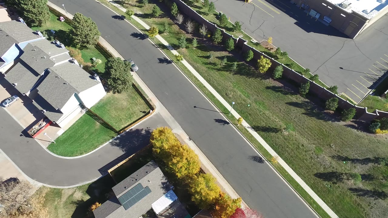 High overhead frame in Hidden Lake, Colorado with a straight road, open grassy lot, sidewalks, rooftops, trees, and no visible people.