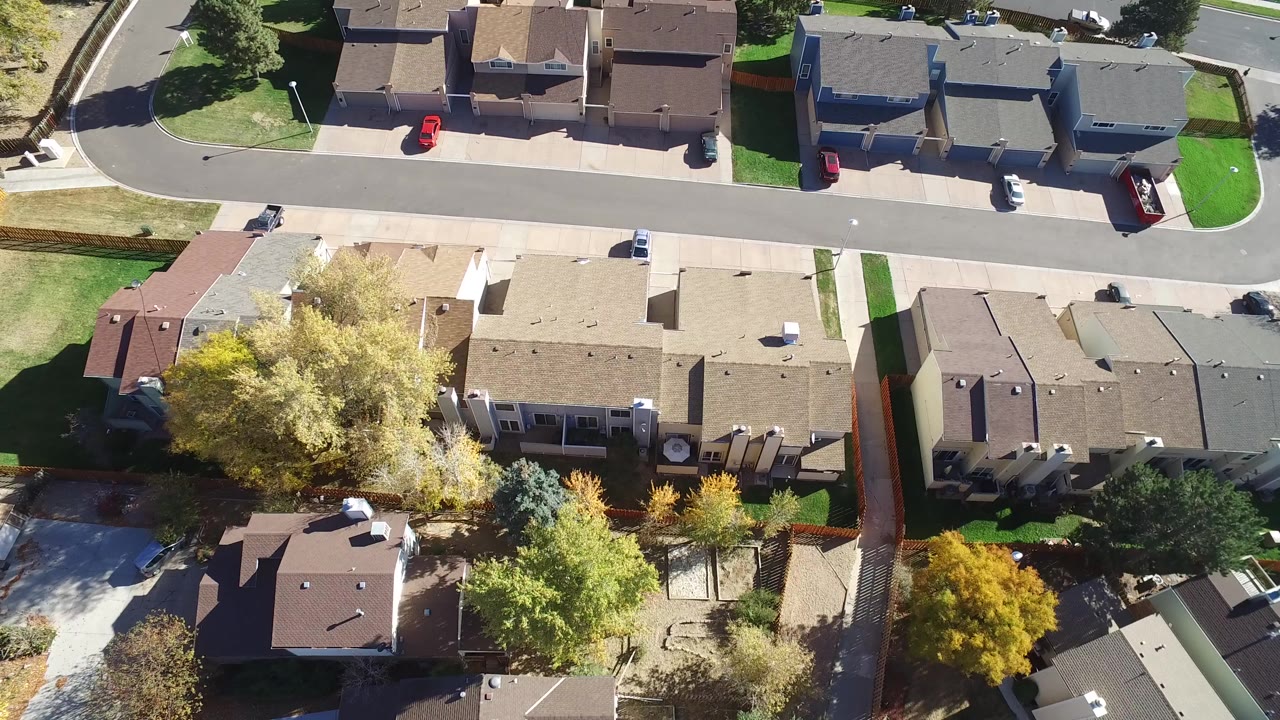 Top-down drone view of residential rooftops, streets, yards, trees, and parked cars.