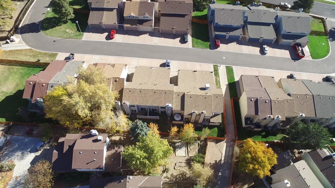Drone-height view over Hidden Lake, Colorado homes with a row of rooftops across the center, a residential street near the top, bright lawns, mature trees, parked cars, and no visible people.