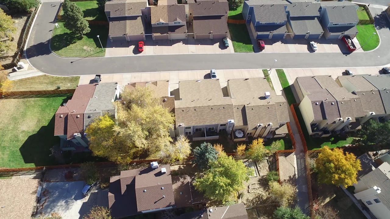 High aerial view over Hidden Lake, Colorado homes with a curved street at the upper left, fenced backyards, bright lawns, mature trees, and no visible people.