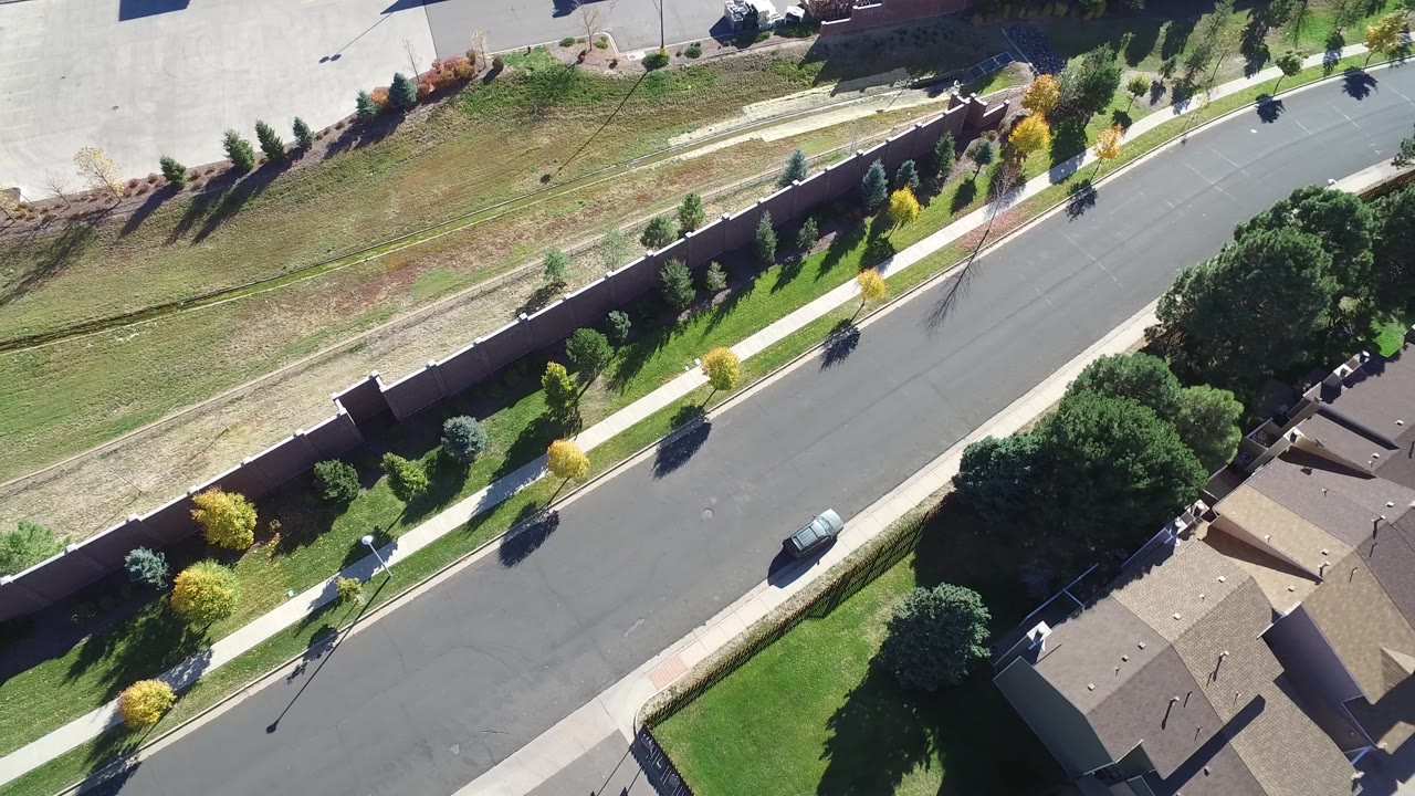 Aerial frame in Hidden Lake, Colorado with the road centered more clearly, a parked vehicle near the curb, yellow and green trees along the sidewalk, and no visible people.
