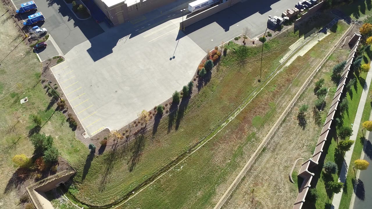 Top-down aerial frame from Hidden Lake, Colorado showing a paved parking area beside a large building, landscaped grass, small trees, a narrow path, and no visible people.