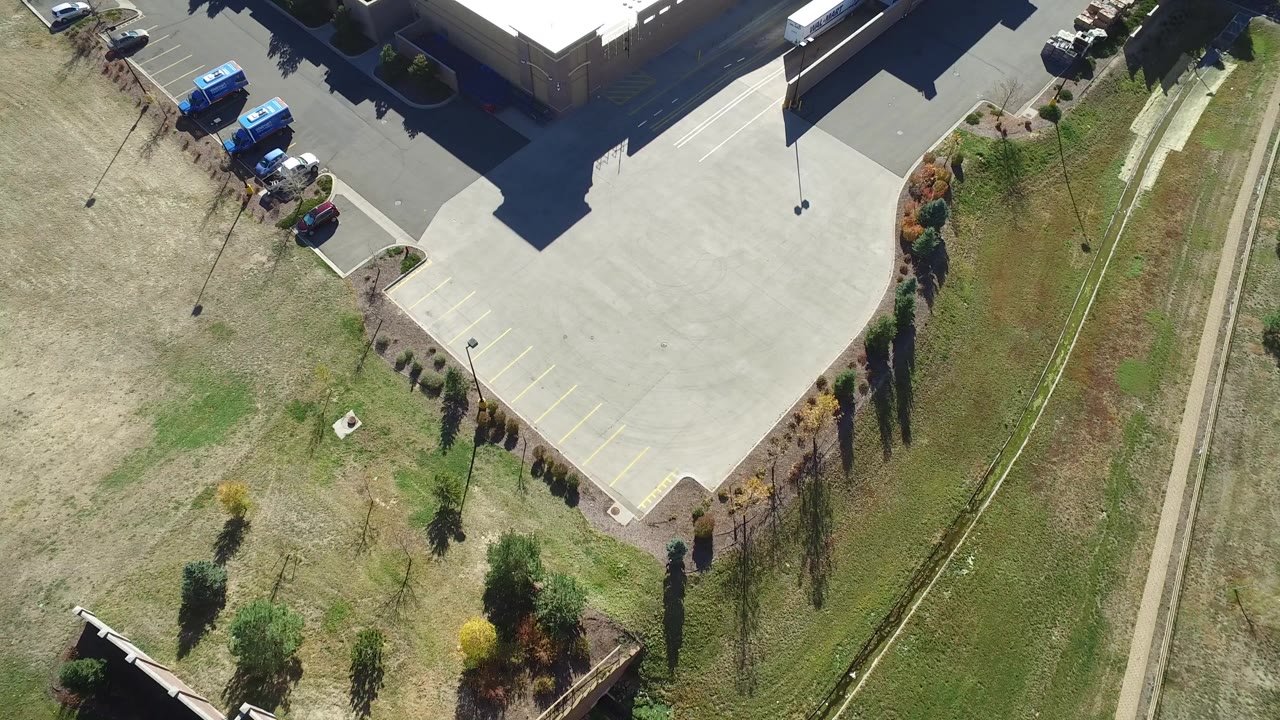 High aerial frame over Hidden Lake, Colorado shows a paved parking-lot corner, dry grass, a building roof edge, parked vehicles, landscaping, and no visible people.