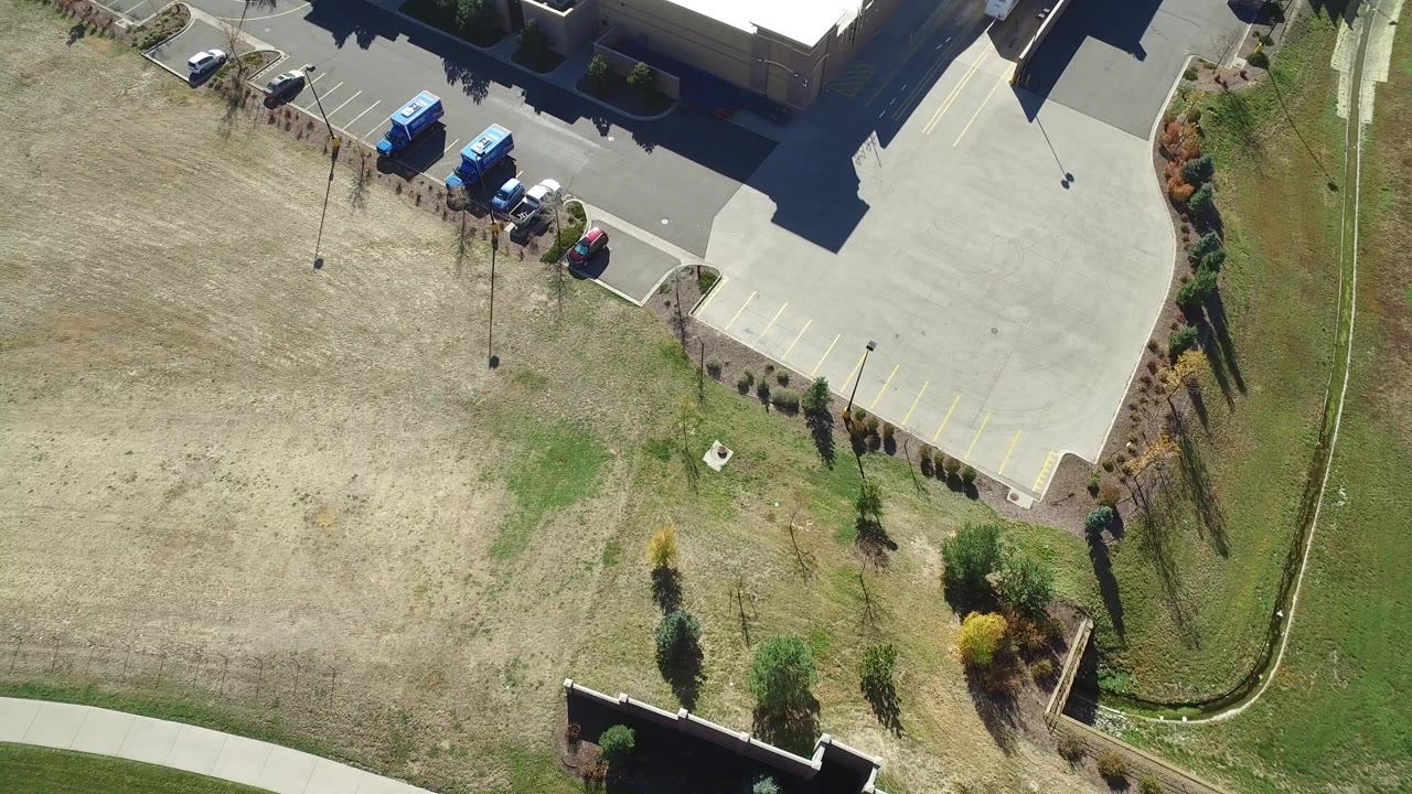Overhead frame in Hidden Lake, Colorado showing a dry grassy field beside a paved parking lot, parked cars, blue trucks, shrubs, a curving sidewalk, and no visible people.