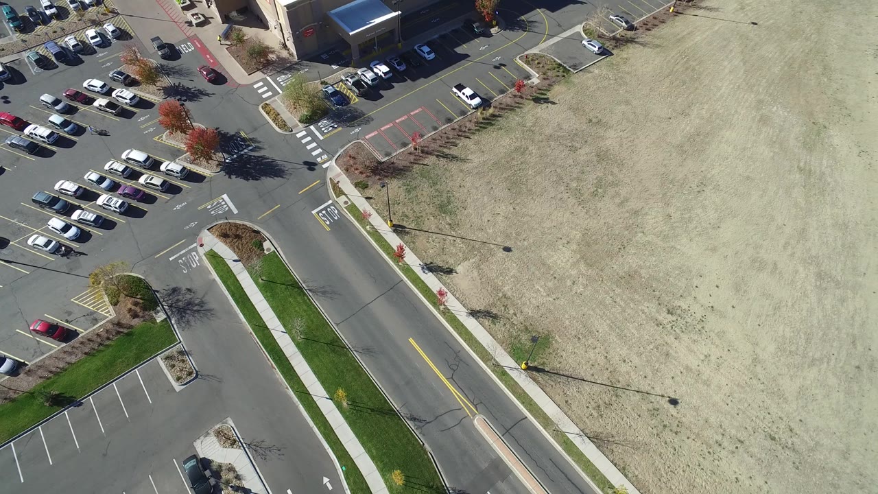 Second frame in Hidden Lake, Colorado with the road running diagonally beside the pale field and clustered parked cars near commercial buildings, with no visible people.