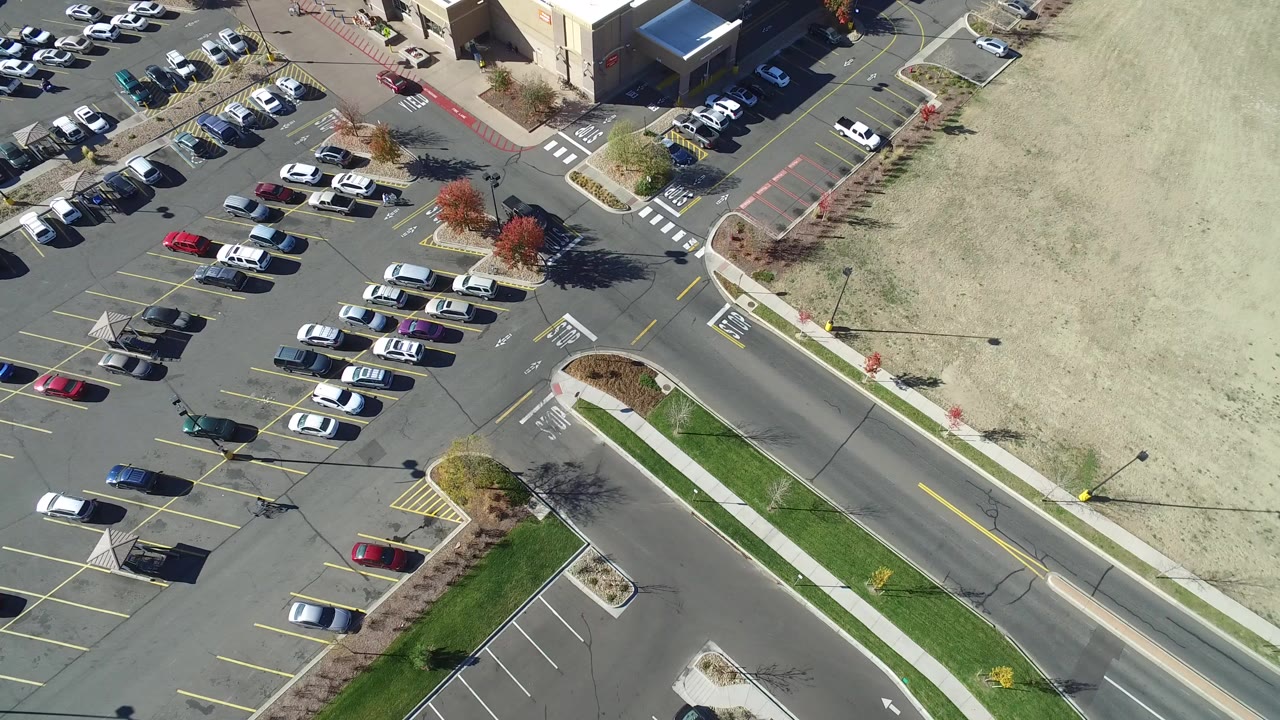 Aerial frame over Hidden Lake, Colorado showing a large parking lot with rows of cars, a road at lower right, sidewalks, grass strips, and part of a department store building.