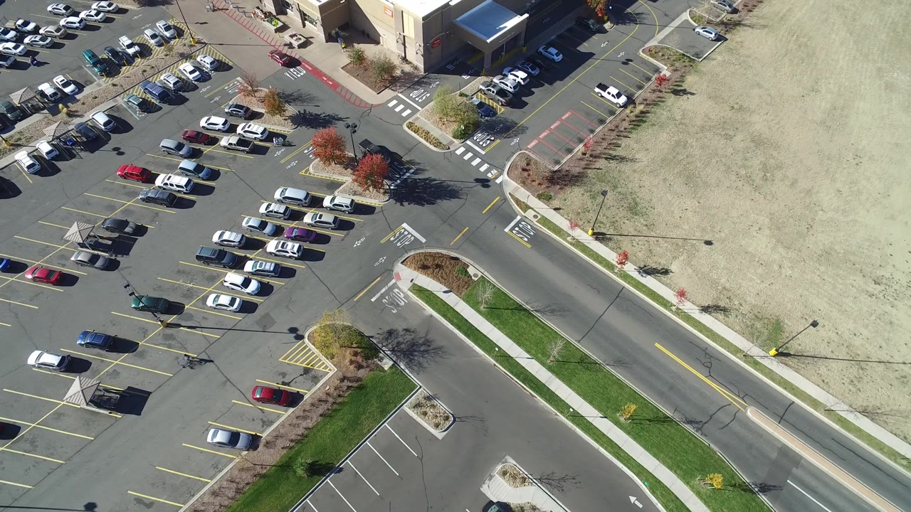 High overhead view in Hidden Lake, Colorado with parked cars arranged in yellow-lined bays, a curving access lane, a road, sidewalks, and a broad bare dirt area beyond the lot.