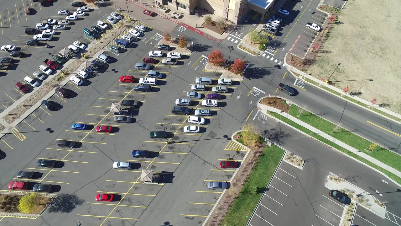 High-angle aerial frame over a Hidden Lake, Colorado parking lot, showing rows of parked cars, yellow stall lines, a road, sidewalks, landscaped islands, and no visible people.