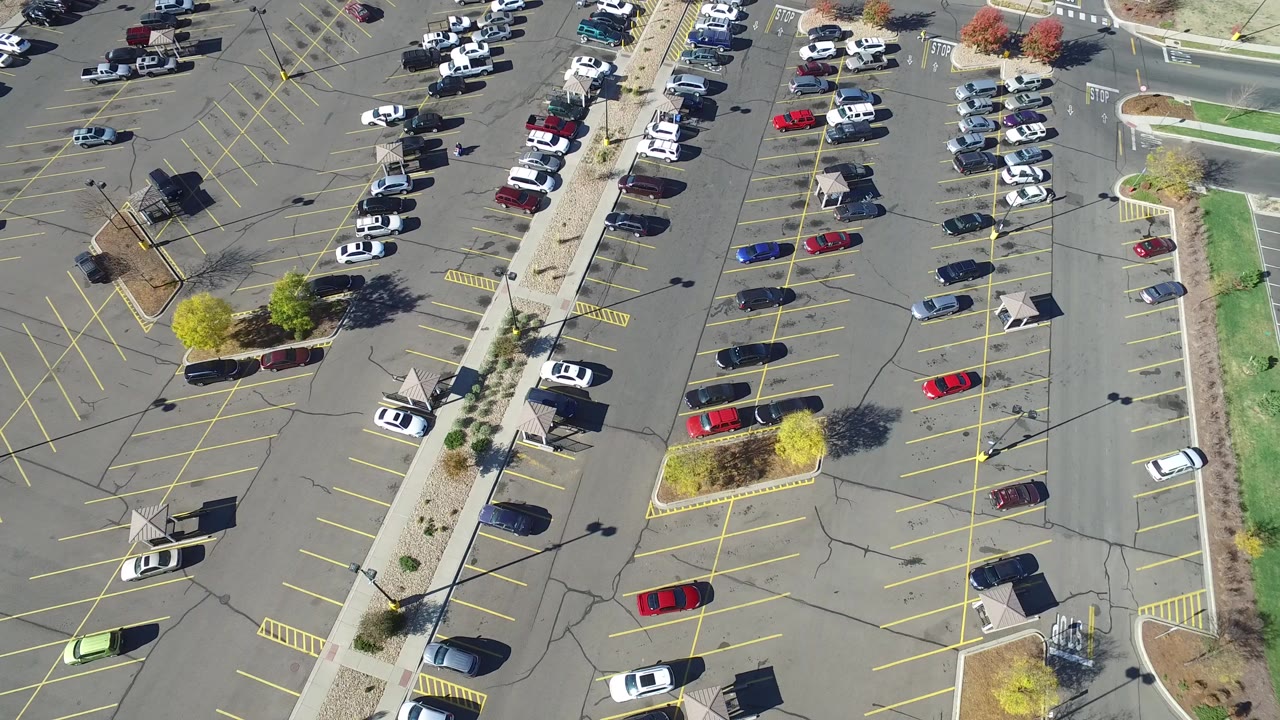 Overhead parking lot with cars, yellow stall lines, sidewalks, and landscaped islands.