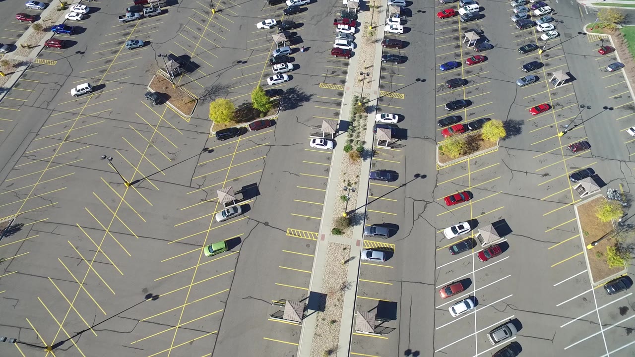 Overhead aerial frame in Hidden Lake, Colorado, showing a broad gray parking lot, diagonal yellow stall markings, a few parked cars near the edges, landscaped islands, and no clearly visible people.