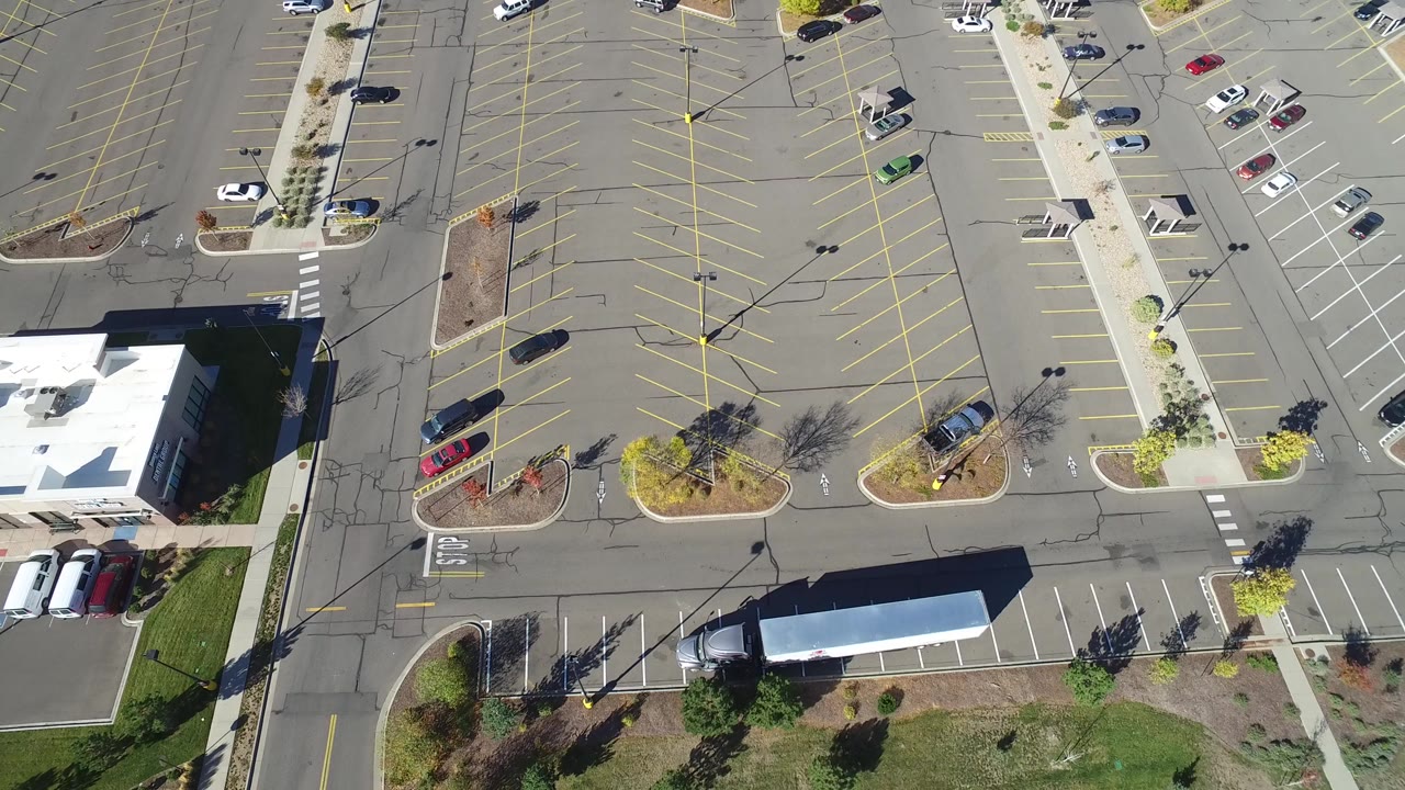 Overhead frame from Hidden Lake, Colorado showing a white-roofed building at lower left, striped parking rows, scattered cars, sidewalks, landscaped islands, and no visible people.